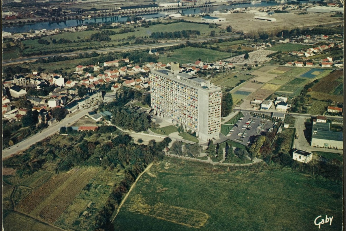 Vue aérienne de la Maison Radieuse, côtés Ouest et Sud ; rue Théodore Brosseaud, le bourg de Rezé avec le château de la Bouvardière et l'hôtel Grignon-Dumoulin, la chapelle Saint-Lupien, le cimetière Saint-Pierre, la route de Pornic, en arrière-plan les villages de la Haute-Ile et de la Basse-Ile, la Loire, l'île de Nantes