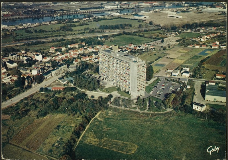 Vue aérienne de la Maison Radieuse, côtés Ouest et Sud ; rue Théodore Brosseaud, le bourg de Rezé avec le château de la Bouvardière et l'hôtel Grignon-Dumoulin, la chapelle Saint-Lupien, le cimetière Saint-Pierre, la route de Pornic, en arrière-plan les villages de la Haute-Ile et de la Basse-Ile, la Loire, l'île de Nantes